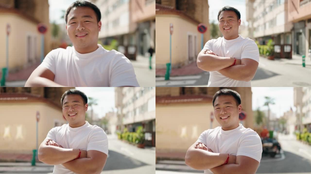 年轻的中国男子微笑着自信地站在街上，双臂交叉手势