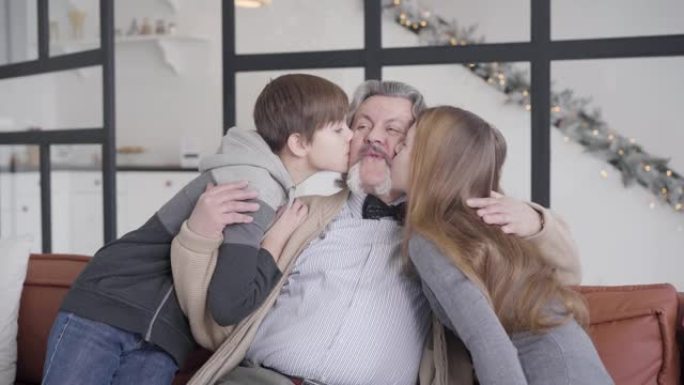 孙子来到祖父那里，坐在沙发上，亲吻脸颊上的老人。圣诞夜在家快乐轻松的多代家庭聚会的肖像。假日概念。