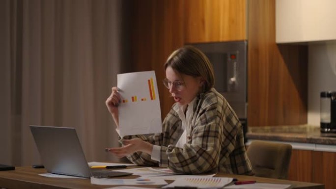 家庭办公室一位戴眼镜的年轻女子坐在厨房里，对着笔记本电脑的摄像头展示图表，进行在线报告。视频电话和会