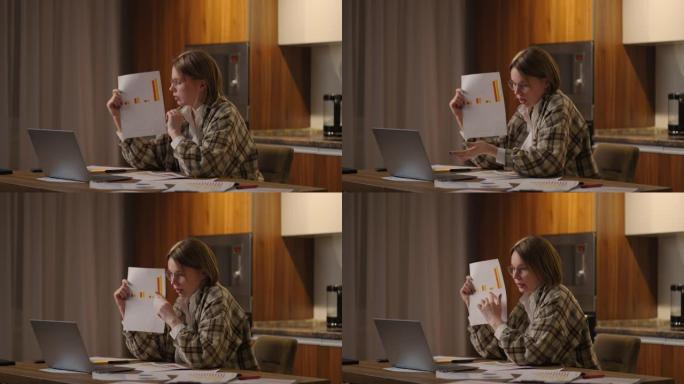 家庭办公室一位戴眼镜的年轻女子坐在厨房里，对着笔记本电脑的摄像头展示图表，进行在线报告。视频电话和会