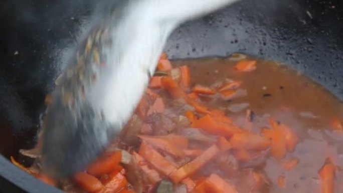 洋葱和胡萝卜在铸铁大锅中油炸。有烟和蒸汽。厨师用开槽的勺子搅动蔬菜。在户外烹饪。特写。