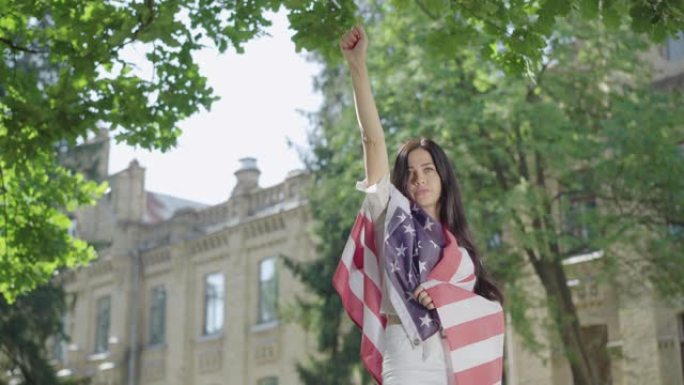 身着美国国旗的黑发美女举起手来。肖像自信迷人的女士显示自由姿态在大学校园。阳光明媚的一天，年轻的大学