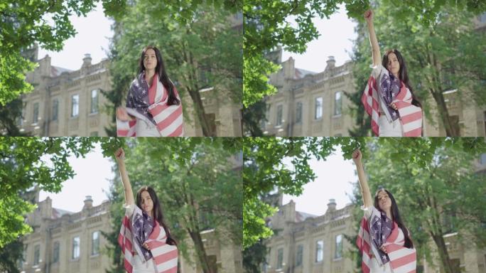 身着美国国旗的黑发美女举起手来。肖像自信迷人的女士显示自由姿态在大学校园。阳光明媚的一天，年轻的大学
