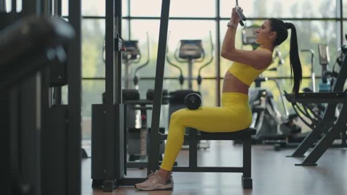 坐在健身房模拟器上的西班牙裔妇女拉一根金属绳，体重会增加背部的肌肉。黑发女人拉模拟器。为背部肌肉模拟