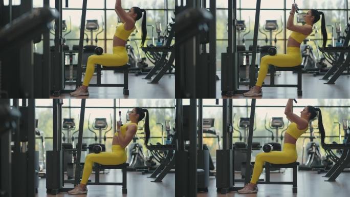 坐在健身房模拟器上的西班牙裔妇女拉一根金属绳，体重会增加背部的肌肉。黑发女人拉模拟器。为背部肌肉模拟