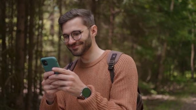 一个背着手机背包的年轻人正在森林地区的一条道路上行走，写信息，在线交流，查看内容，微笑，喜欢散步。