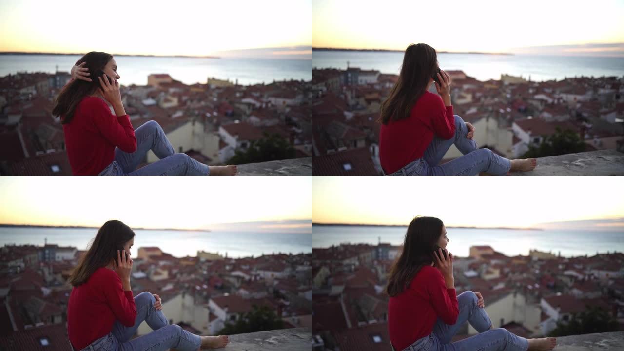 年轻女子坐在壁架上，看着大海，同时在日落时用手机