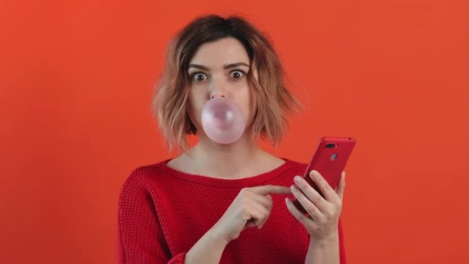 年轻性感女孩的肖像红色毛衣在社交网络背景中翻阅红色智能手机，口香糖和红色背景上微笑的膨胀气泡。乐事。