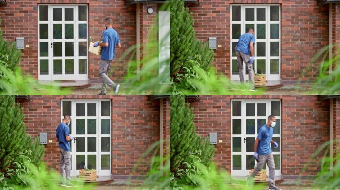 戴着防护口罩和手套的年轻人在家庭家门口留下了装有新鲜农产品的木箱