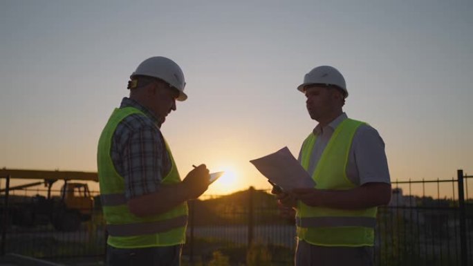 两个建筑商的手的肖像。建筑商握手建筑商在建房背景上。在太阳的背景下，两个穿着绿色信号背心的男人握手的