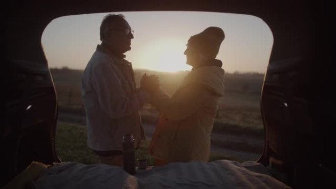 两个老年人在公路旅行中露营。站在大型汽车后备箱旁边互相温暖的手
