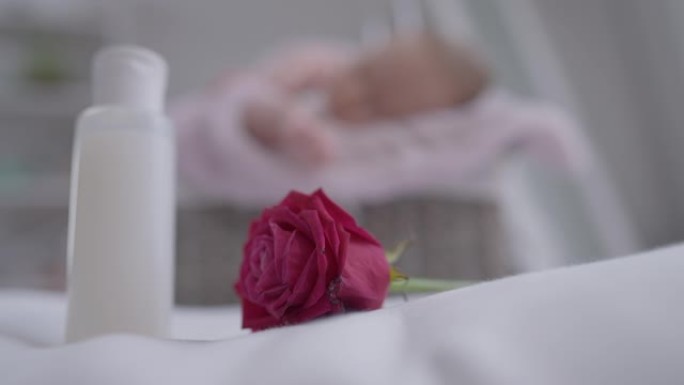 红色美丽的玫瑰花躺在室内，女性手拿婴儿爽身瓶，模糊的婴儿在背景下睡觉。白人母亲在家照顾小女儿。慢动作