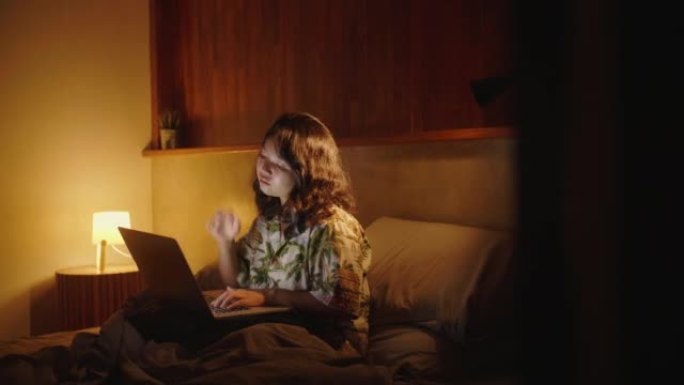 亚洲年轻女子晚上坐在床上，在卧室里使用笔记本电脑。