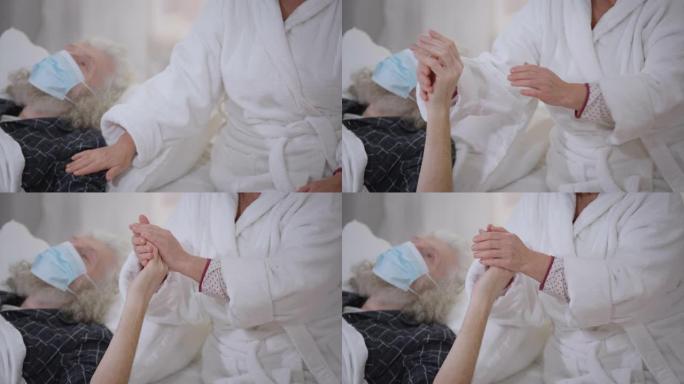 面目全非的高级妻子爱抚着生病的丈夫的手躺在床上，戴着Covid-19的口罩。充满爱心的白人妇女抚摸着