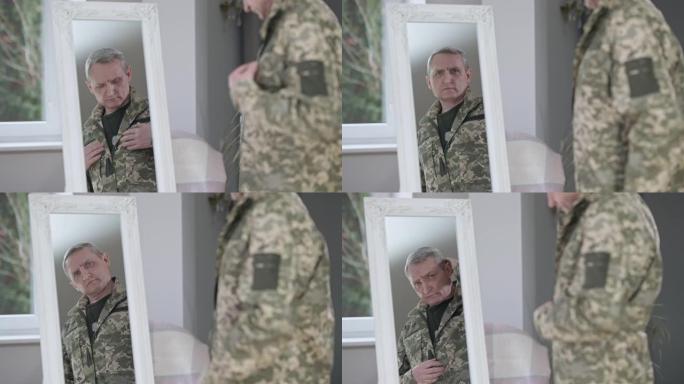 自信英俊的中年男子在室内调整军装的镜子中映照。一幅严肃的白人士兵在家伪装的照片。步兵服务。