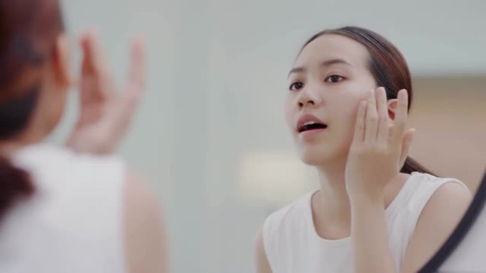 亚洲女孩看着镜子，在脸上涂抹面部保湿霜，在化妆前进行面霜处理。
