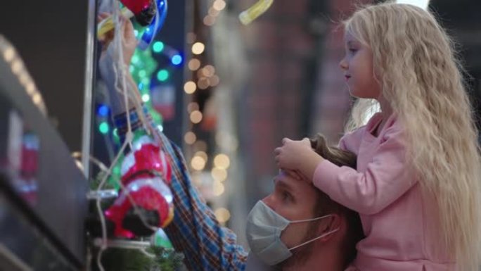在大流行期间，坐在购物中心肩膀上的父女在慢动作中选择房子的花环和圣诞节的圣诞树。