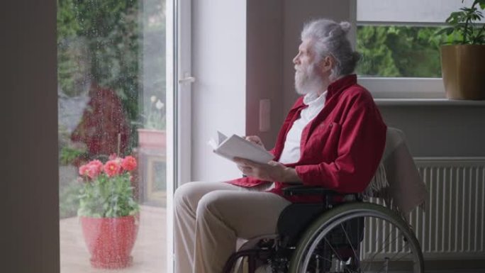 坐在轮椅上的高级残疾人在雨天看着窗外。聪明的残疾高加索退休人员的肖像在家中享受文学作品。慢动作。