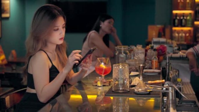 年轻的亚洲人在手机上玩耍，感到悲伤，晚上在酒吧和餐馆喝鸡尾酒。