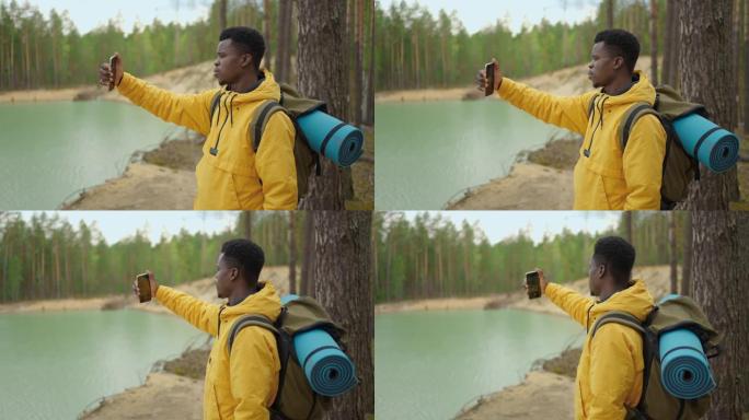 一个黑人正走在山顶上，在智能手机上输入消息和照片。他肩上有一个大背包。在山里徒步旅行。山顶