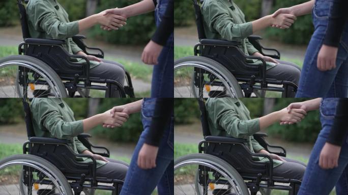 侧视图无法识别的残疾高加索女孩在慢动作中问候朋友握手。坐在轮椅上的截瘫少年在夏季公园的户外会见朋友。