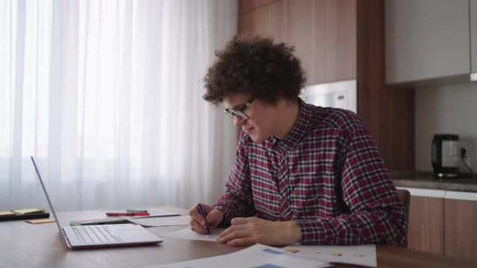 卷发男学生戴眼镜的迷人小男孩正在家里学习，用笔记本电脑打字，用笔记本电脑写字。大学生使用笔记本电脑观