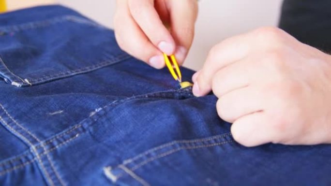 一个人用镊子从胶带上取下保护膜，用双面胶带将接缝处撕裂的牛仔裤粘合起来。无能的人修理破衣服，侧视图。