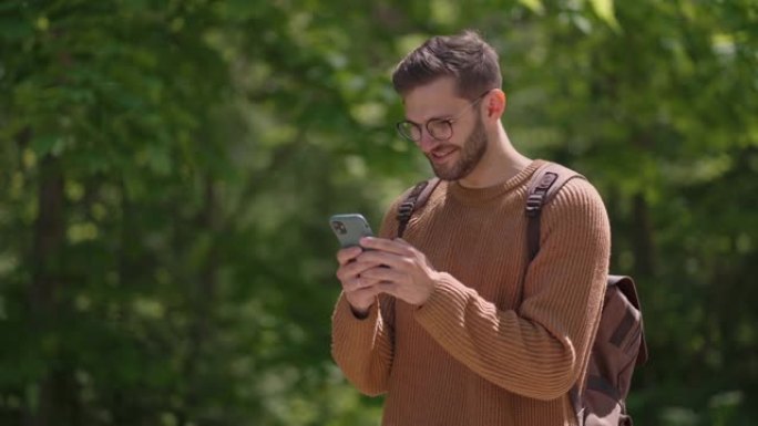 一名男性游客带着手机背包在森林地区写信息，在线交流，查看内容，微笑，享受散步。