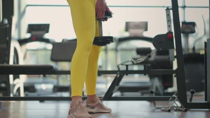 年轻的西班牙裔黑发女子穿着黄色运动服蹲在健身房里拿着哑铃