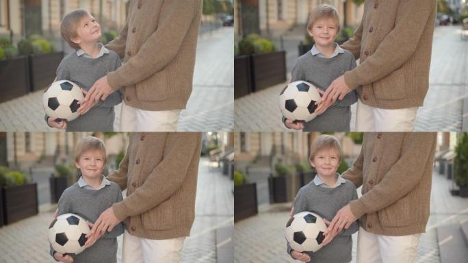 迷人的小男孩拿着足球，在阳光下与面目全非的老人合影。快乐微笑的高加索孙子在城市街道上与祖父一起享受周