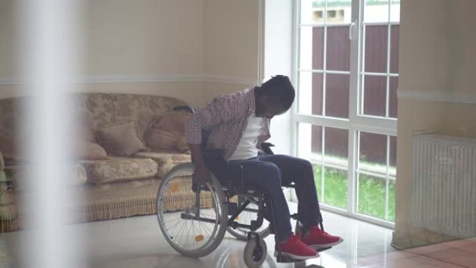 快乐的非洲裔美国残疾人在室内轮椅上慢动作跳舞。宽幅肖像微笑的英俊瘫痪的家伙在客厅里在家中打手势成功。