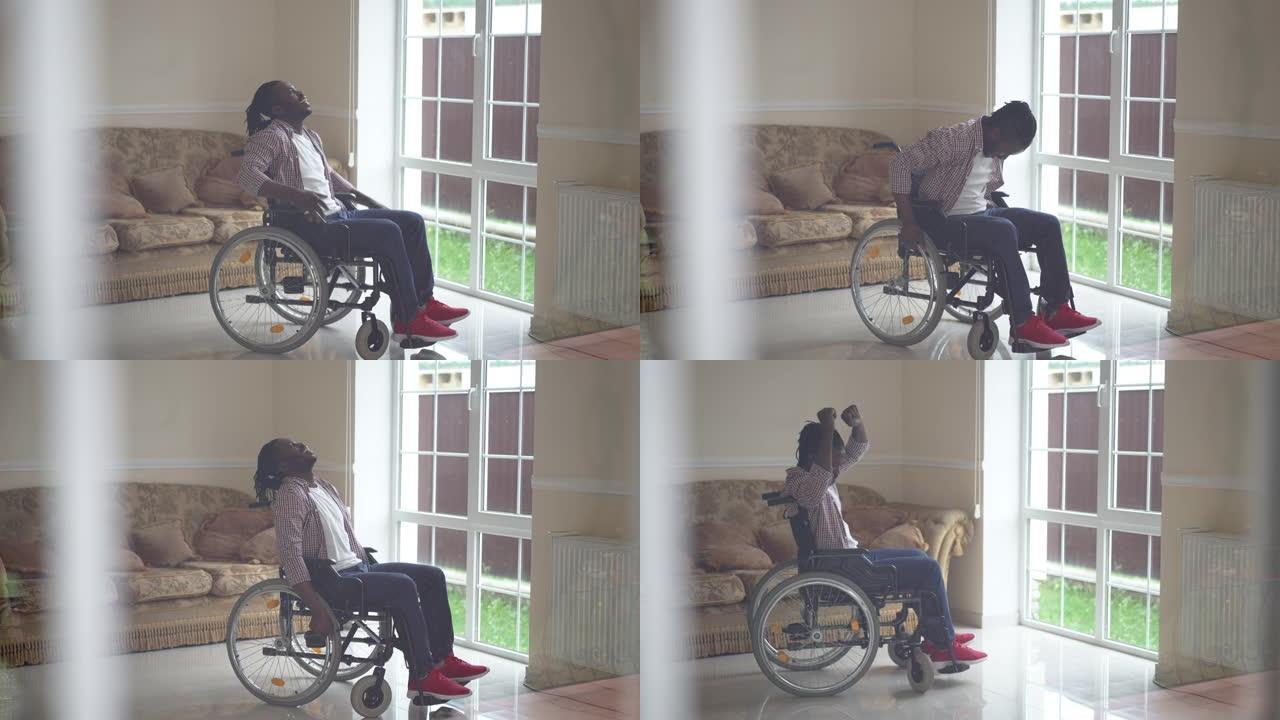 快乐的非洲裔美国残疾人在室内轮椅上慢动作跳舞。宽幅肖像微笑的英俊瘫痪的家伙在客厅里在家中打手势成功。
