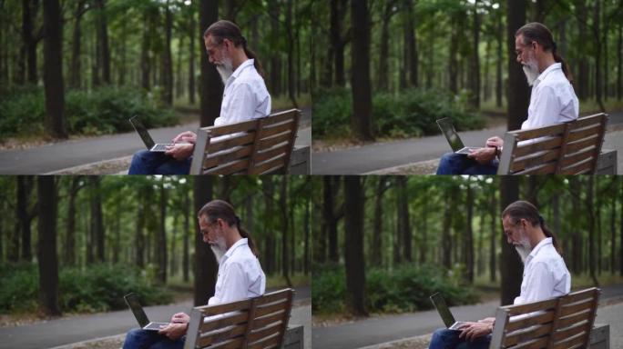 一个成年男子在公园长椅上留着灰色胡须，与笔记本电脑一起工作。4k慢动作视频。