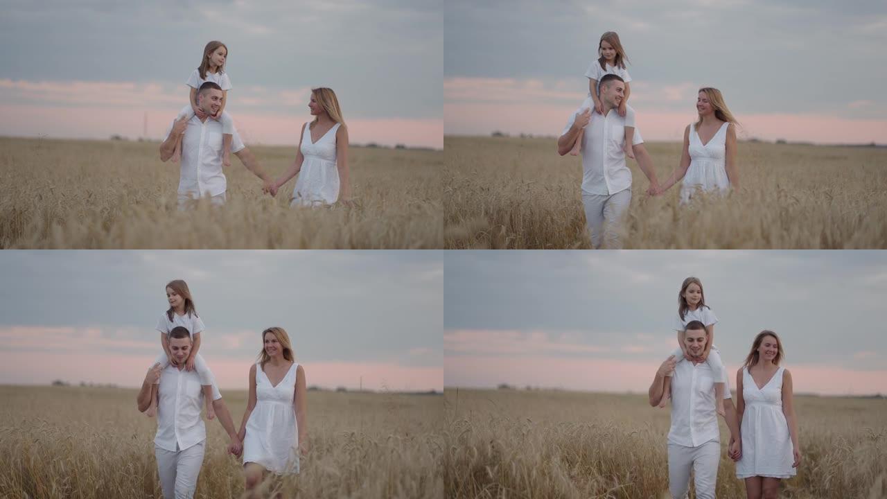 一对年轻的父母和女童手牵着手，在日落时分穿过麦田。幸福的家庭在大麦草地上慢跑，一起享受大自然。慢动作