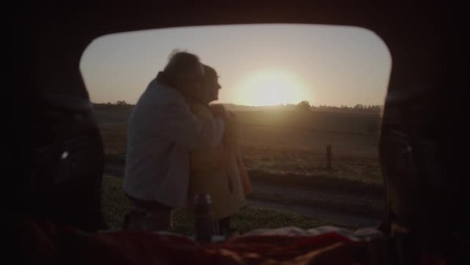 两个老年人在公路旅行中露营。站在大型汽车后备箱旁边互相拥抱
