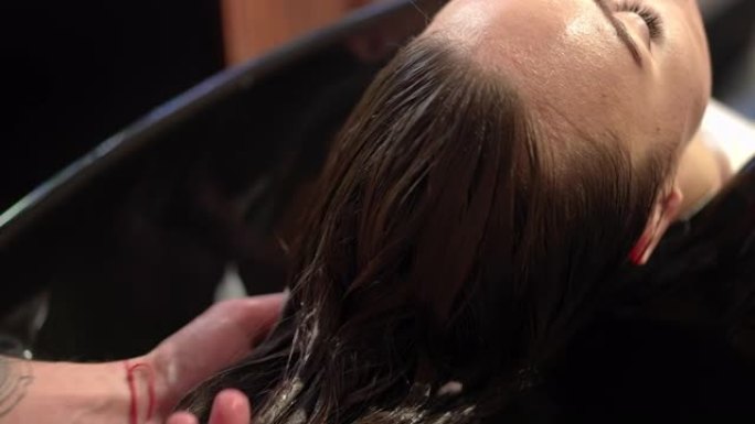 在美发沙龙中，特写男性手在女性客户的棕色长发上涂抹护发素。无法识别的白人男子用乳液油在慢动作中抚摸头