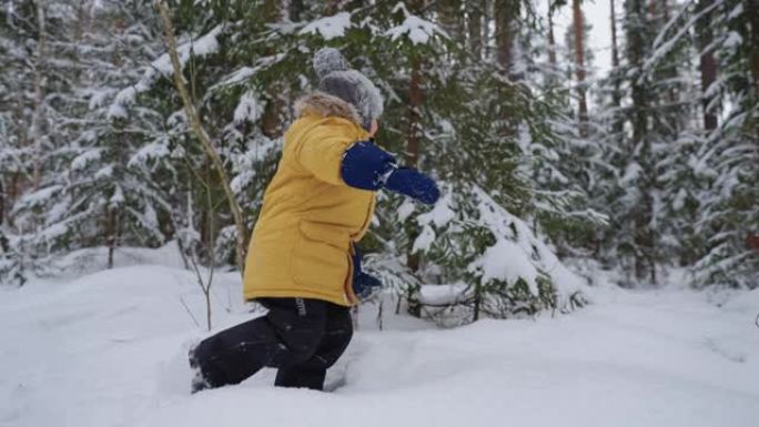 一个穿着黄色外套的男孩穿过深雪，研究冬季森林，冬季行走，并以慢动作穿过雪林。儿童自由环境的概念