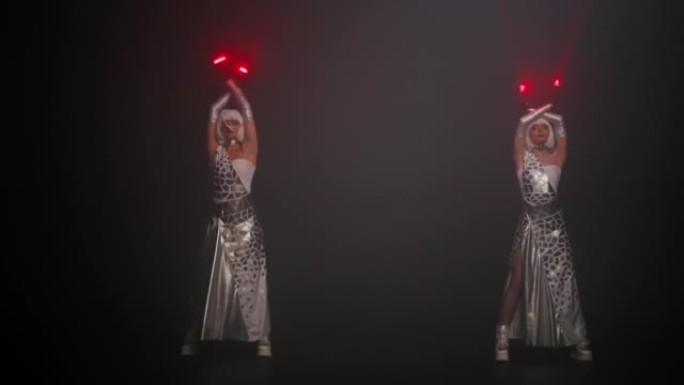 两名穿着网络服装的妇女在舞台上的雾中表演，在手套上打开红色霓虹灯。穿着未来派服装与激光束共舞的才华横