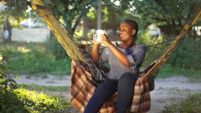 微笑的大码女人用相机拍照，欣赏户外夏日自然之美。坐在吊床上微笑着看着远处的才华横溢的非洲裔美国摄影师