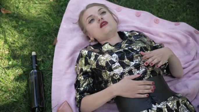 俯视图醉酒的年轻女子躺在公园绿色夏季草坪上的毯子上唱歌。空酒瓶一边。悲伤醉酒的高加索千禧一代女士在户