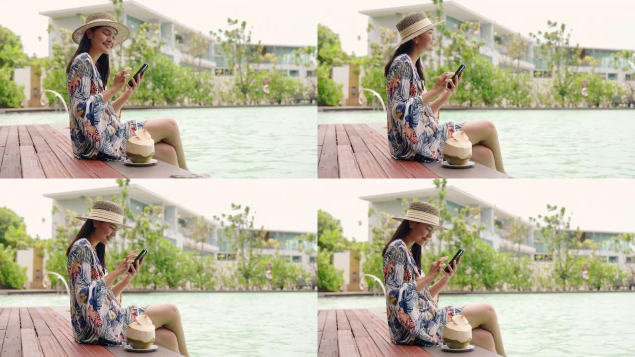女人坐在游泳池边喝椰子水放松玩手机。
