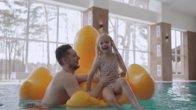 快乐的小女孩在游泳池里笑，坐在有趣的充气火烈鸟里，爸爸抱着他的女儿