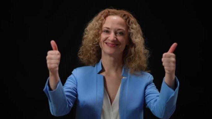 快乐的女人在黑色背景的相机闪光灯中竖起大拇指。微笑自信的高加索优雅政客看着相机打手势的肖像。成功理念