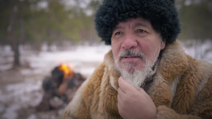 老自信的土著男子抚摸着灰色的胡须，面带微笑。老人坐在冬季森林中的肖像，背景下慢动作燃烧着火。生活方式
