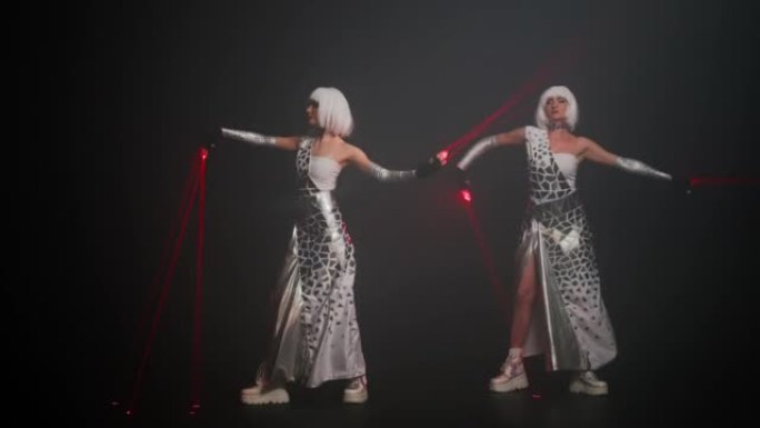 两个穿着未来派服装的苗条美女，带着激光束在舞台上跳舞变换的地方。自信的有才华的白人表演者在黑色背景下