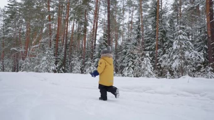 一个3-4岁的小男孩在冬季森林中奔跑，穿着黄色外套从后面慢动作观看。冬季乐趣和积极娱乐的概念，自由和