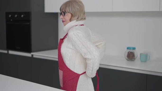 穿着眼镜和白色毛衣系围裙的华丽高级女性站在家里的厨房室内。自信的退休人员烹饪食物享受周末的肖像。生活