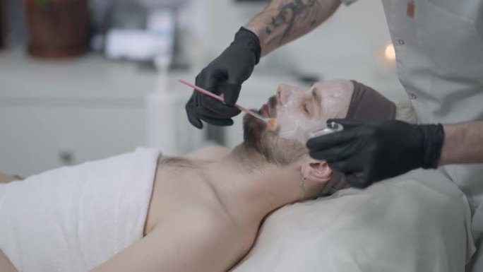 侧视年轻男子闭着眼睛躺在纹身的男性美容治疗师用刷子在慢动作中涂抹保湿面霜。大胡子的白人客户喜欢清洗程