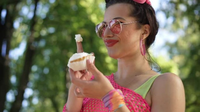 年轻的coquette高加索女人穿着复古的衣服，在慢动作中品尝甜美美味的甜甜圈奶油，站在夏日公园大笑