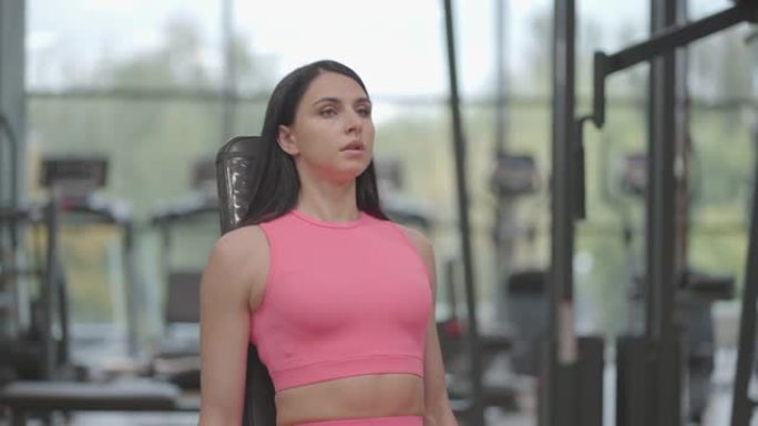 一个穿着粉红色西装的黑发女人在健身房训练肩膀时向侧面举起哑铃。坐式肩部和手臂长凳练习